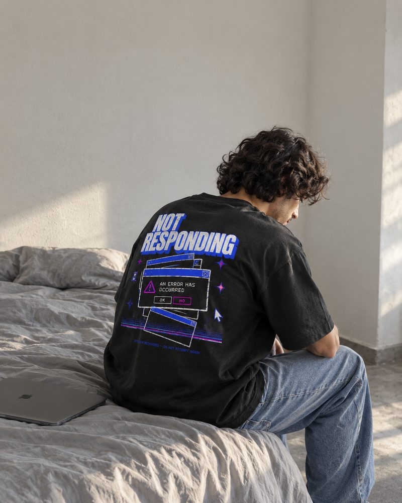 Not Responding tee, back print, seated on the edge of a bed beside a closed laptop